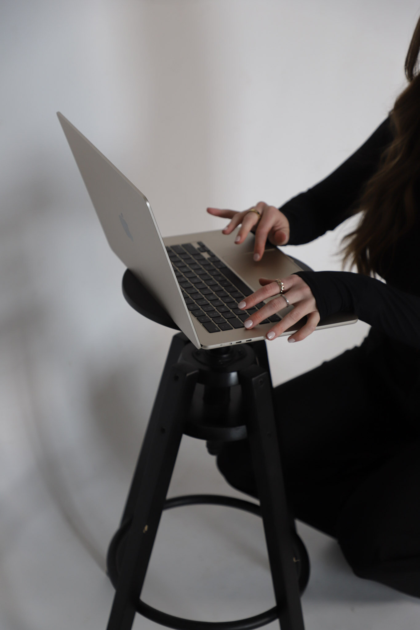 Women in black, typing on a laptop on a stool.